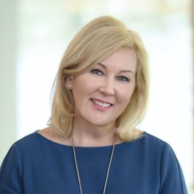 Smiling light-skinned, female-presenting individual with medium length blonde hair wearing a blue shirt on a grey backdrop.