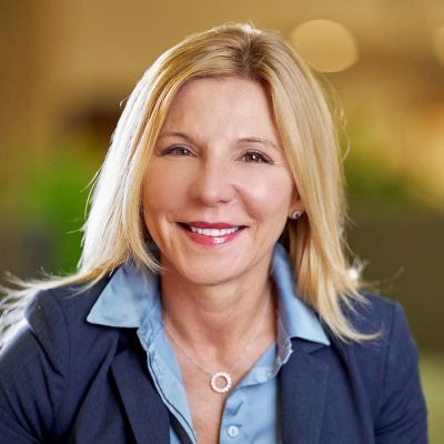 Smiling light-skinned, female-presenting individual with long blonde hair wearing a blue suit on a brown backdrop.