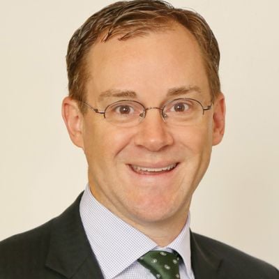 Smiling light-skinned, male-presenting individual with short hair wearing a dark suit on a beige backdrop.