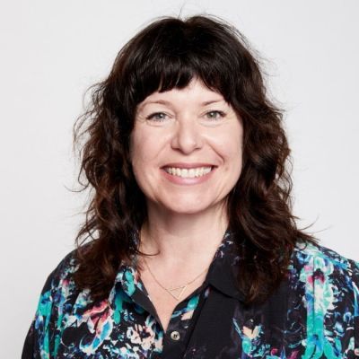 A smiling light-skinned female with dark hair wearing a patterned shirt against a light gray background