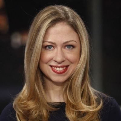 A smiling light-skinned female with long blonde hair wearing a dark shirt against a dark background