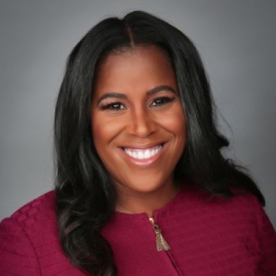 A smiling darker-skinned female with long black hair wearing a cranberry colored shirt against a gray background