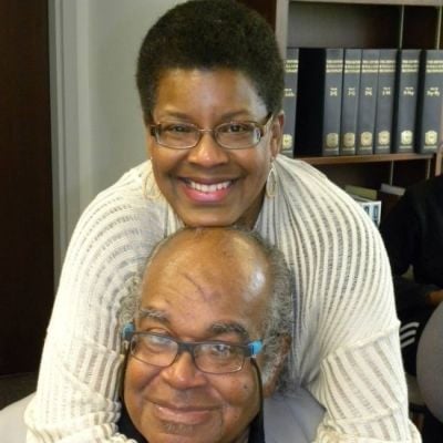 Two smiling darker-skinned individuals wearing glasses with a bookshelf in the background