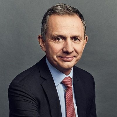 Smiling light-skinned, male-presenting individual with short grey hair wearing a dark suit on a grey backdrop.