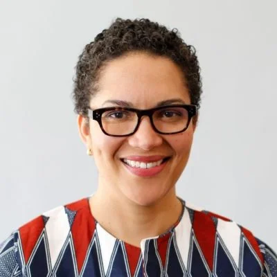 A smiling darker-skinned female with short dark brown hair wearing glasses and a patterned shirt against a light gray background