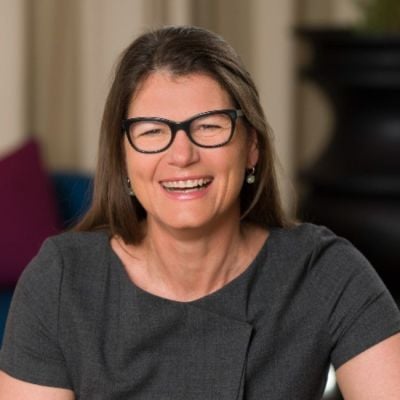 Smiling light-skinned, female-presenting individual with long dark hair wearing a dark shirt on a brown backdrop.