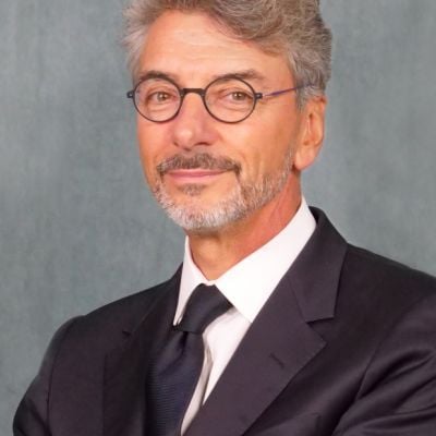 Smiling light-skinned, male-presenting individual with short grey hair wearing a dark suit on a grey backdrop.