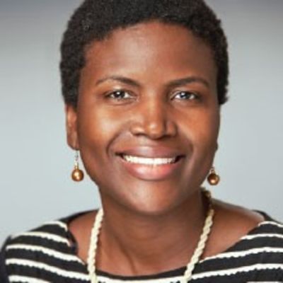 Smiling darl-skinned, female-presenting individual with short dark hair wearing a black and white striped shirt on a grey backdrop.