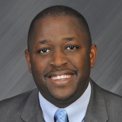 A smiling dark-skinned male wearing a gray suit and tie against a gray striped background