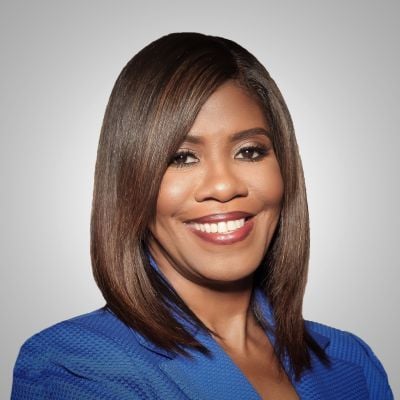 Smiling dark-skinned, female-presenting individual with medium dark hair wearing a blue jacket on a grey backdrop.