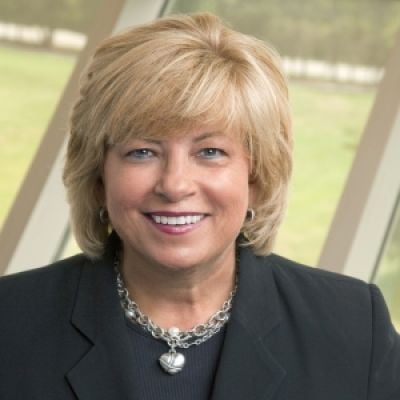 A smiling light-skinned female with blonde hair wearing a black suit against an outdoor background