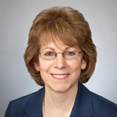 A smiling light-skinned female with light brown hair wearing a navy suit against a light gray background