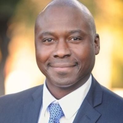 Smiling dark-skinned, male-presenting individual with short hair wearing a dark suit on an outdoor backdrop.