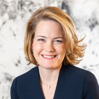A smiling light-skinned female-presenting individual wearing a navy suit jacket with short blonde hair against a marbled background