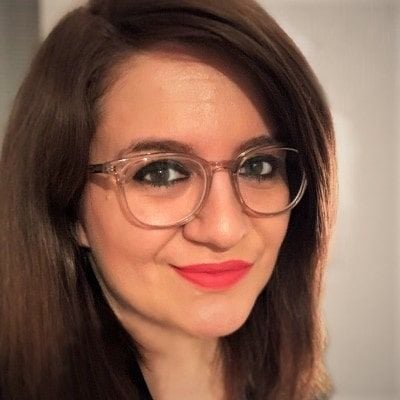 A smiling light-skinned female with dark brown hair and glasses against an ambiguous background