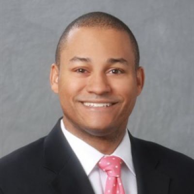 A smiling darker-skinned male wearing a dark suit against a gray gradient background