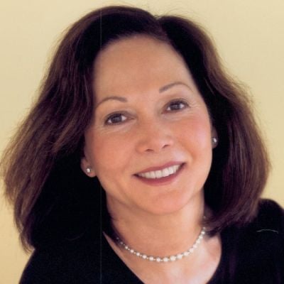 A smiling light-skinned female with dark brown hair wearing a black shirt against a beige background