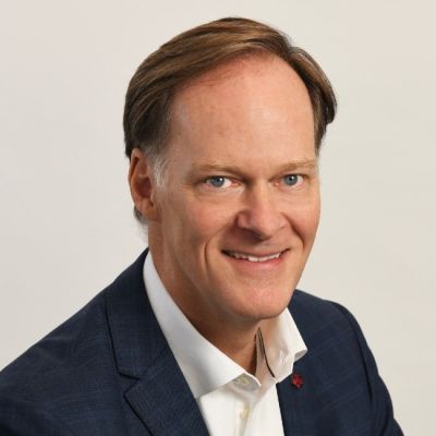 A smiling light-skinned male-presenting person with light brown hair wearing a navy suit jacket and a white button-up against a light gray background
