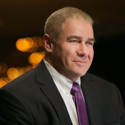 A smiling light-skinned male with short light brown and gray hair wearing a black suit and tie against a dark background