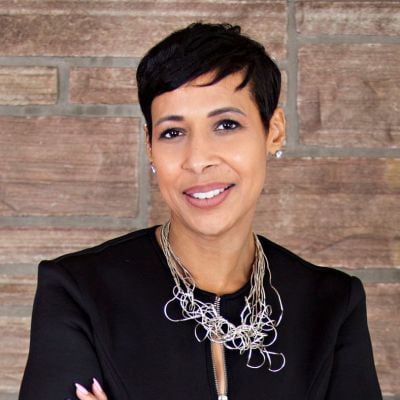 Smiling female-presenting individual with short dark hair wearing a black jacket on a brick backdrop.