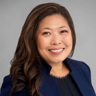 A smiling female-presenting individual of Asian heritage wearing a navy suit jacket against a gray background