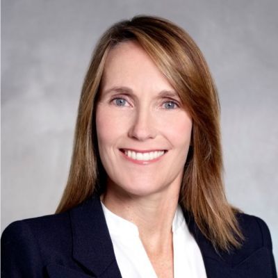 Smiling light-skinned, female-presenting individual with long red hair wearing a dark suit on a grey backdrop.