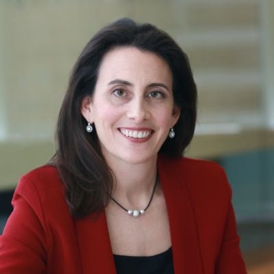 Smiling light-skinned, female-presenting individual with long dark hair wearing a red jacket on a brown backdrop.