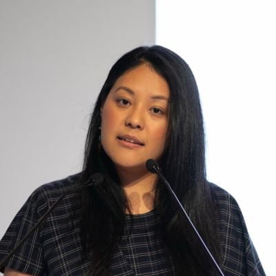 A female-presenting person with long black hair wearing a dark gray and black patterned shirt speaking in front of a microphone