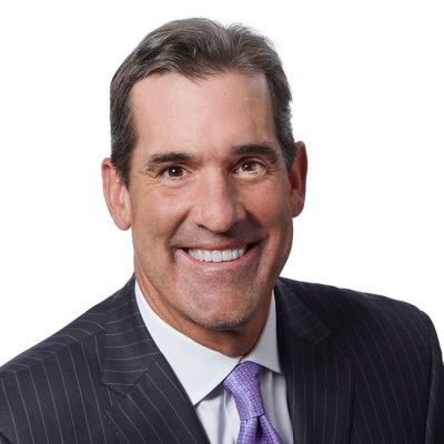 Smiling light-skinned, male-presenting individual with short dark hair wearing a dark suit on a white backdrop.