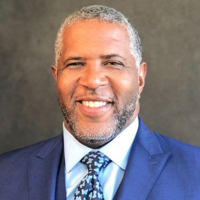 Smiling dark-skinned, male-presenting individual with short grey hair wearing a blue suit on a grey backdrop.