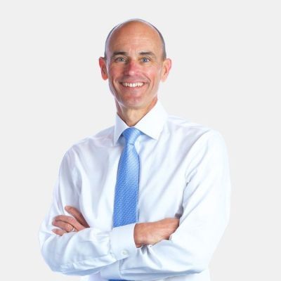 Smiling light-skinned, male-presenting individual with short grey hair wearing a white shirt, blue tie on a beige backdrop.