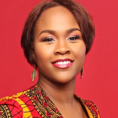 Smiling dark-skinned, female-presenting individual with short dark hair wearing a printed shirt on a red backdrop.