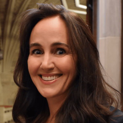 Smiling light-skinned, female-presenting individual with long dark hair wearing a dark shirt on a brown backdrop.