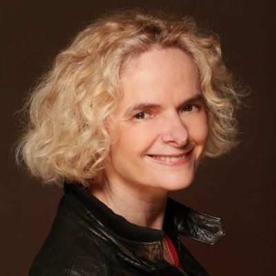 A smiling light-skinned female with short blonde hair wearing a dark black jacket against a dark brown background