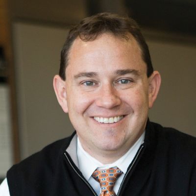 A smiling light-skinned male with short dark brown hair wearing a black jacket and tie against a brown background