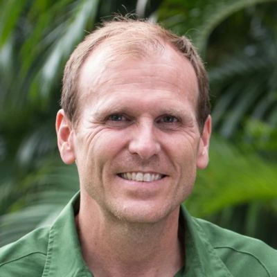 A smiling light-skinned male-presenting individual with light brown hair wearing a green shirt against an outdoor background