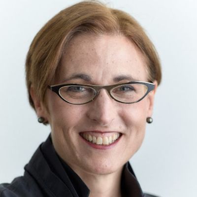Smiling light-skinned, female-presenting individual with short red hair wearing a dark shirt on a white backdrop.