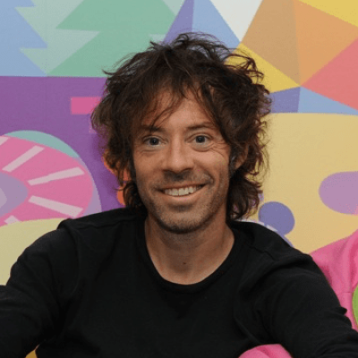 A smiling light-skinned male with long dark brown hair wearing a black shirt against an abstract colorful background