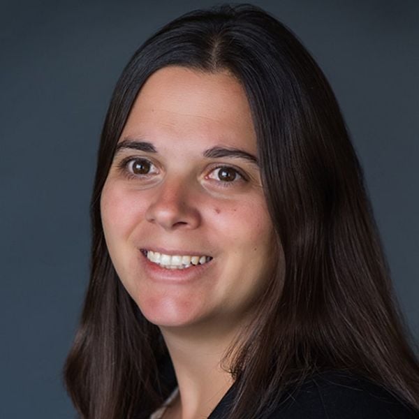 A smiling light-skinned female with shoulder length dark brown hair wearing a dark shirt against a dark gray background