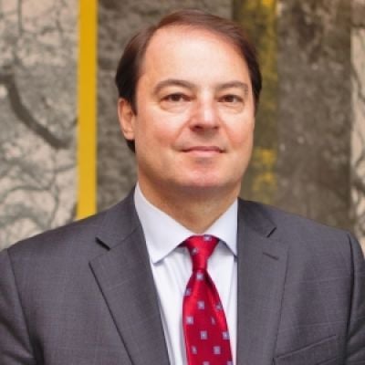 A light-skinned male with brown hair wearing a gray suit and red tie against an abstract background