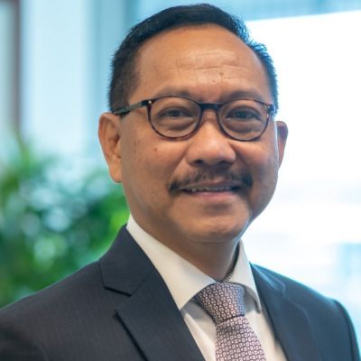 A smiling male of Asian heritage wearing a dark suit and tie and glasses against a blurred background