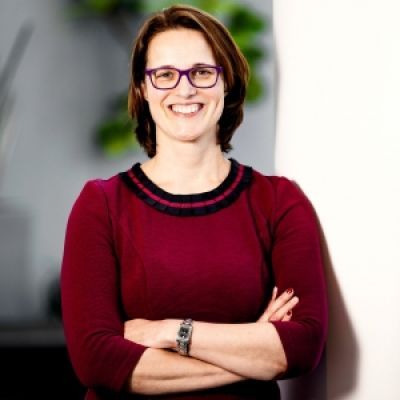 A smiling light-skinned female with short brown hair wearing a dark red sweater and glasses against a blurred background