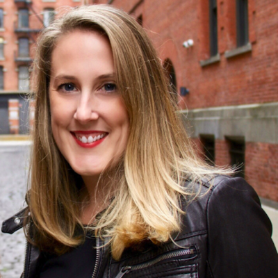 A smiling light-skinned female with blonde hair wearing a black leather jacket against a background of brick houses