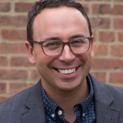 A smiling lighter-skinned male wearing glasses and a dark gray suit jacket over a button-up shirt against a brick wall