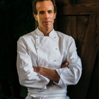 A light-skinned male with dark brown hair wearing a chef's coat against a dark background