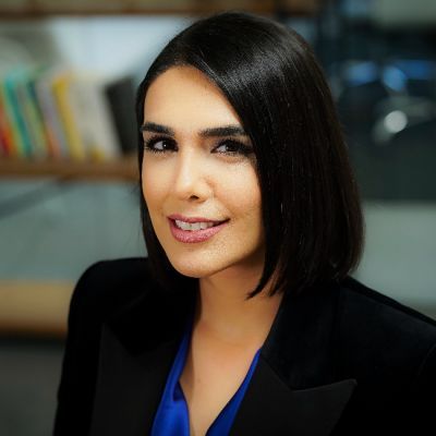 A smiling light-skinned female with short black hair wearing a black suit jacket over a blue shirt against a blurred backdrop