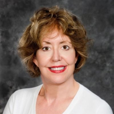 A smiling light-skinned female with shorter blonder hair wearing a white shirt against a gray marbled background