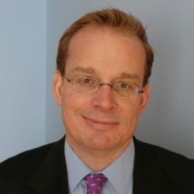 A smiling light-skinned male with short brown hair wearing glasses and a dark suit and tie against a light blue background