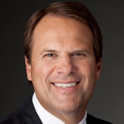 A smiling light-skinned male with dark brown hair wearing a suit against a dark gray background