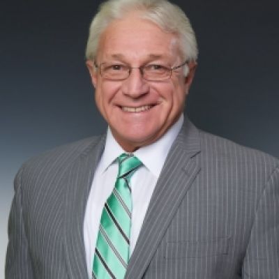 A smiling light-skinned male with white hair wearing glasses and a gray suit and tie against a dark gray background